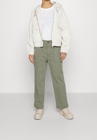 Beige kordjacka med huva, vit t-shirt, olivgröna lösa byxor och beige kraftiga stövlar. Texturerad tyg och avslappnad siluett.