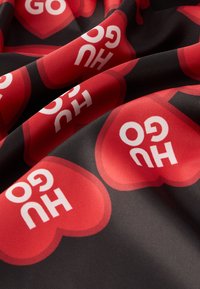 Crvena i crna tkanina s velikim, preklapajućim crvenim srcima i bijelim natpisom "GO HUG". Materijal izgleda gladak s blagim sjajem.