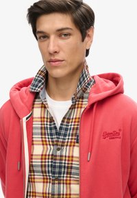 Rode hoodie met een rits aan de voorkant, witte voering en grijze hardware, gelaagd over een meerkleurig geruit overhemd met bruine knopen.