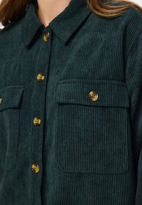 Σκούρο πράσινο μπουφάν κοτλέ με μεγάλες μπροστινές τσέπες και κουμπιά από χελώνα, που φοριέται από ένα άτομο με ανοιχτά καστανά μαλλιά.