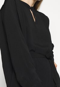 Schwarzes langärmeliges Kleid mit einem Schlitz-Ausschnitt und einem Stoffband in der Taille. Verfügt über strukturierte vertikale Streifen an den Ärmeln.