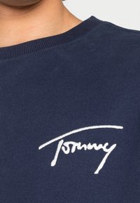 Närbild av en marinblå crewneck-tröja med en vit broderad "Tommy"-logotyp på bröstet, bärs av en person.