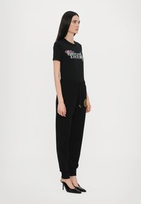 Črna majica s cvetličnim potiskom "COUTURE", kombinirana s črnimi trenirkami z zlatim emblemom in črnimi visokimi petami. Preprost dizajn.