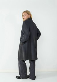 Dunkelgrauer Wollmantel mit Kragen, lockerer Passform, seitlichen Taschen und langen Ärmeln; kombiniert mit weit geschnittenen grauen Hosen und schwarzen Schuhen.