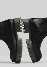 Scarpe alte in pelle nera con suola grigia spessa e textured, caratterizzata da sezioni ritagliate e una finitura liscia. Presentano pannelli laterali elastici.