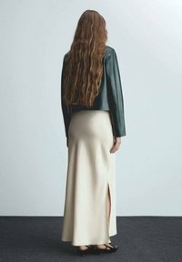 Černá kožená zkrácená bunda, smetanová dlouhá sukně s bočním rozparkem, modelka s dlouhými vlnitými vlasy, stojící na šedém koberci na světlém pozadí.