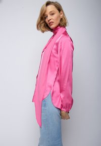 Jasnoróżowa satynowa bluzka z wysokim dekoltem i długimi rękawami, o zwiewnym kroju z bocznymi rozcięciami, w połączeniu z jasnym dżinsowym materiałem.