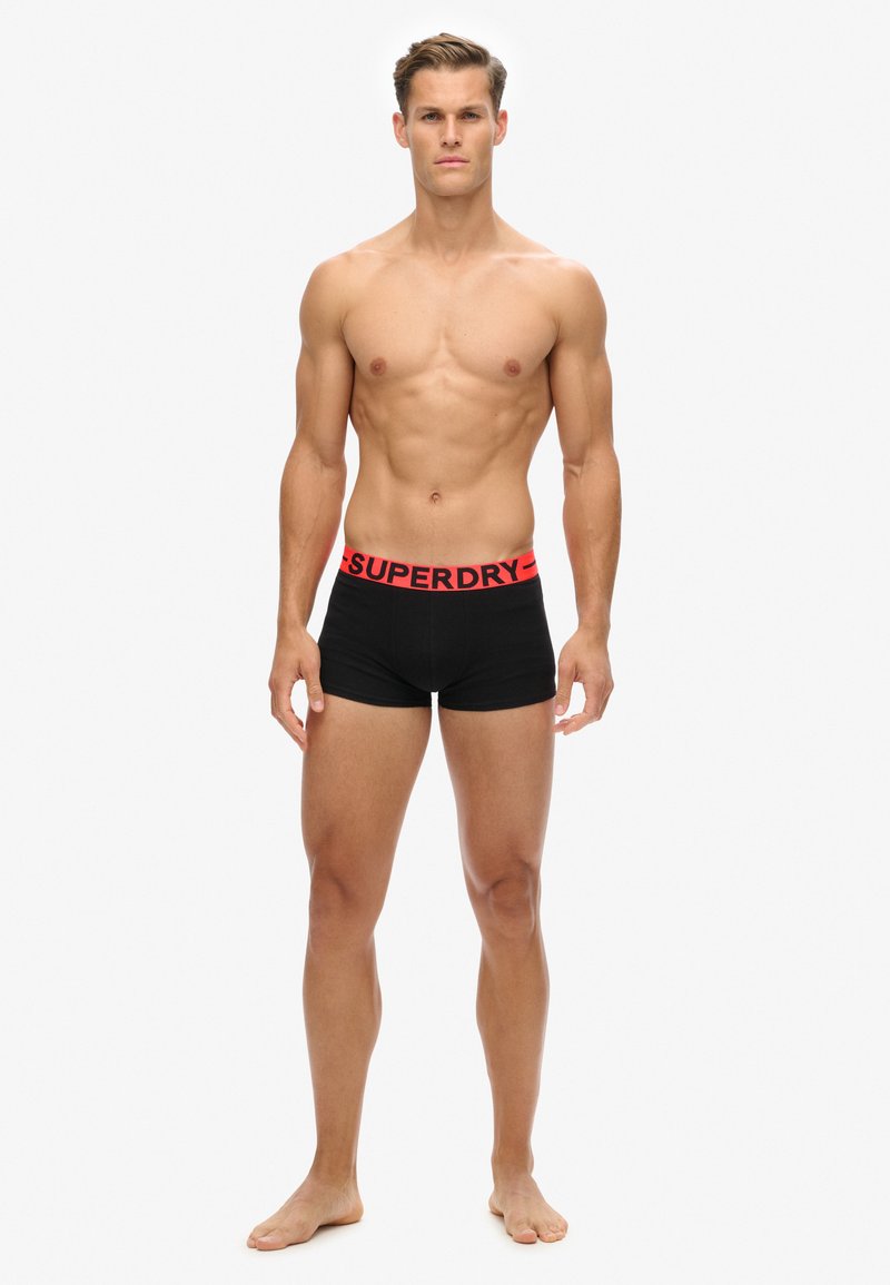 Schwarze Boxer-Briefs mit einem breiten roten Bund, der mit "SUPERDRY" beschriftet ist. Der Stoff wirkt glatt und dehnbar, und das Design ist körperbetont.