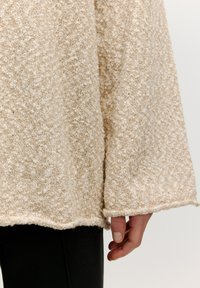 Beiger strukturierter Pullover mit lockerer Passform, weiten Ärmeln und rohem Saum. Der Stoff wirkt weich und hat eine leicht flauschige Oberfläche.