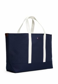 Borsa a tracolla blu navy realizzata in tessuto resistente, con manici bianchi, un accento in pelle marrone e un piccolo logo sulla parte anteriore.
