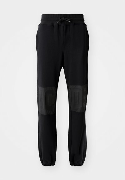 Sorte joggebukser med ribbestrikket liv, snøring og elastiske mansjetter. Har skinnende svarte paneler på lårene med embossed skrift.