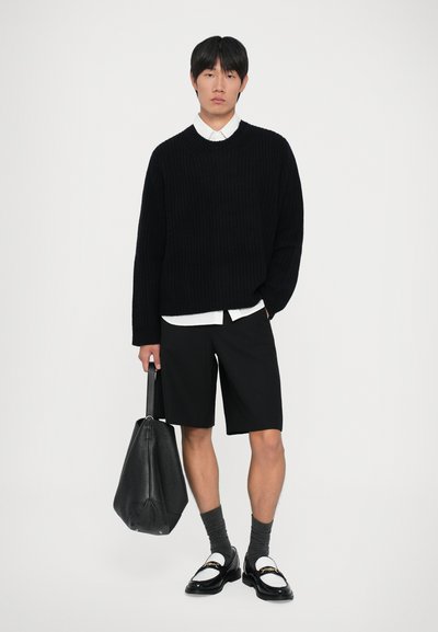Giovane uomo che indossa un maglione di maglia nera, una camicia bianca, pantaloni corti neri, mocassini bianco e neri con calze grigie, mentre tiene una grande borsa a tracolla di pelle nera.