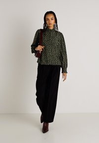 Πράσινη φλοράλ μπλούζα με ψηλό λαιμό, μακρύ μανίκι και χαλαρή γραμμή, συνδυασμένη με μαύρα παντελόνια ψιλό καβαλό και καφέ μποτάκια με τακούνι.