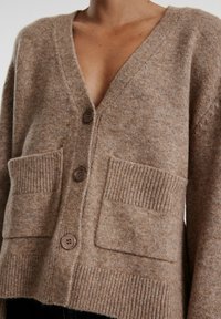 Cardigan de lana marrón con escote en V, que cuenta con dos bolsillos delanteros y tres botones. Tejido texturizado, corte casual y puños acanalados.