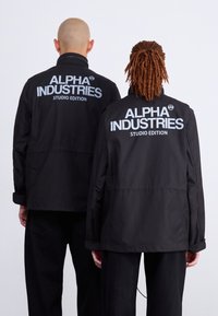 To personer står med ryggen til, iført sorte jakker med teksten "ALPHA INDUSTRIES STUDIO EDITION" trykt i hvidt på ryggen.
