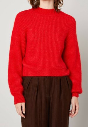 Femme portant un pull en maille rouge vif à manches longues associé à un pantalon plissé ample de couleur marron foncé.
