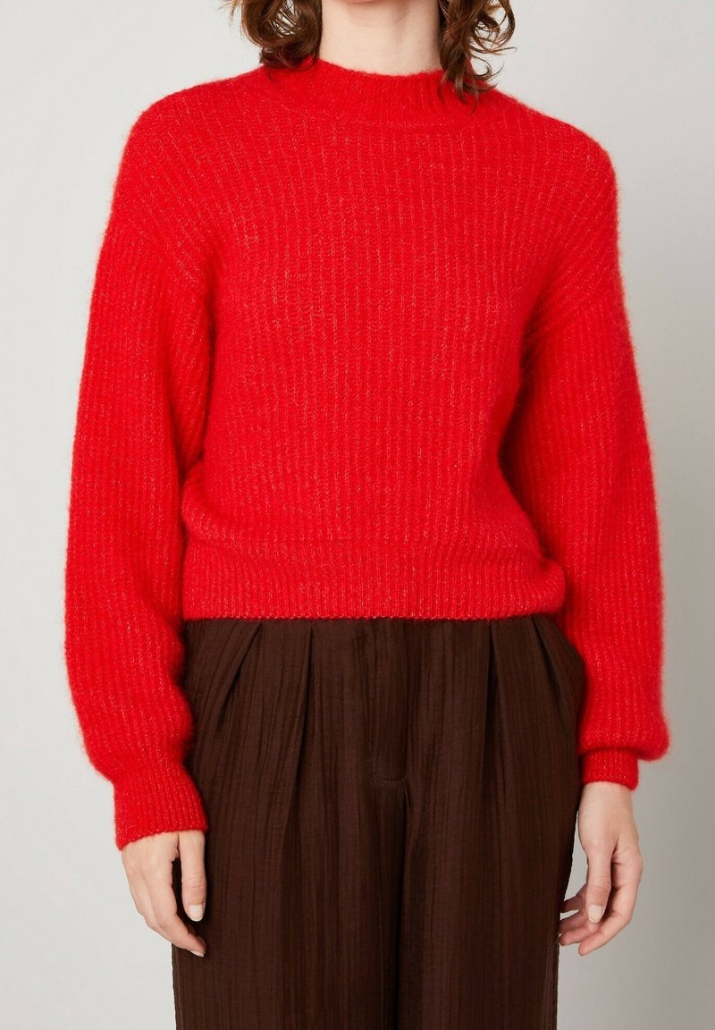 Femme portant un pull en maille rouge vif à manches longues associé à un pantalon plissé ample de couleur marron foncé.