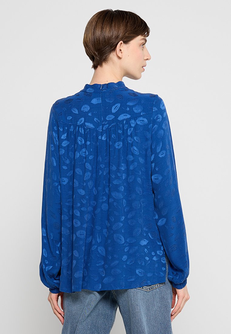 Frau mit kurzem braunem Haar, die eine langärmlige blaue Bluse mit einem dezenten Lippenmuster trägt, von hinten betrachtet.