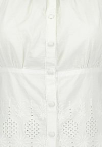 Blusa bianca in cotone con chiusura a bottoni, corpino arricciato e intricati ricami floreali lungo l'orlo con motivi a intaglio.