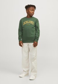 Felpa verde con testo giallo "JACK&JONES" e logo, polsini e orlo a coste, abbinata a pantaloni bianchi e sneakers bianchi.
