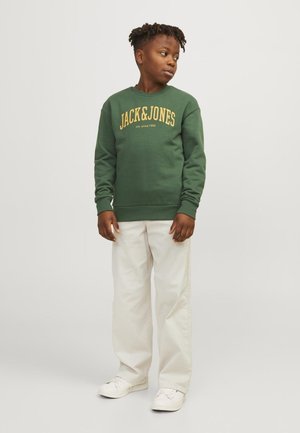 Felpa verde con testo giallo "JACK&JONES" e logo, polsini e orlo a coste, abbinata a pantaloni bianchi e sneakers bianchi.