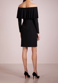 Svart off-shoulder klänning med ett flerskiktat volangdesign, figurnära silhuett, långa ärmar och zip-detalj i ryggen. Spetsiga svarta klackar.