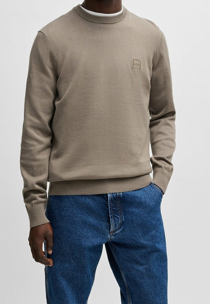Homme portant un pull marron clair à manches longues avec un petit logo carré et un jean denim bleu, debout avec une main dans la poche.