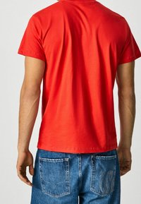 Camiseta de algodón roja de manga corta y cuello redondo, mostrada desde la espalda. Combinada con jeans de mezclilla azul con costura en los bolsillos traseros.