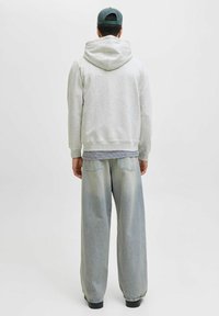 Hellgrauer Kapuzenpullover mit großer Kapuze, über einem gestreiften Hemd getragen. Locker sitzende, hellblaue Jeans mit ausgewaschenem Muster und zwei Gesäßtaschen. Dunkle Schuhe.