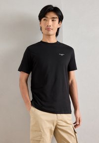 Melnas kokvilnas T-krekls ar apaļu kakla izgriezumu, īsām piedurknēm un nelielu baltu logo uz krūtīm. Gluda tekstūra, brīva piegriezuma.