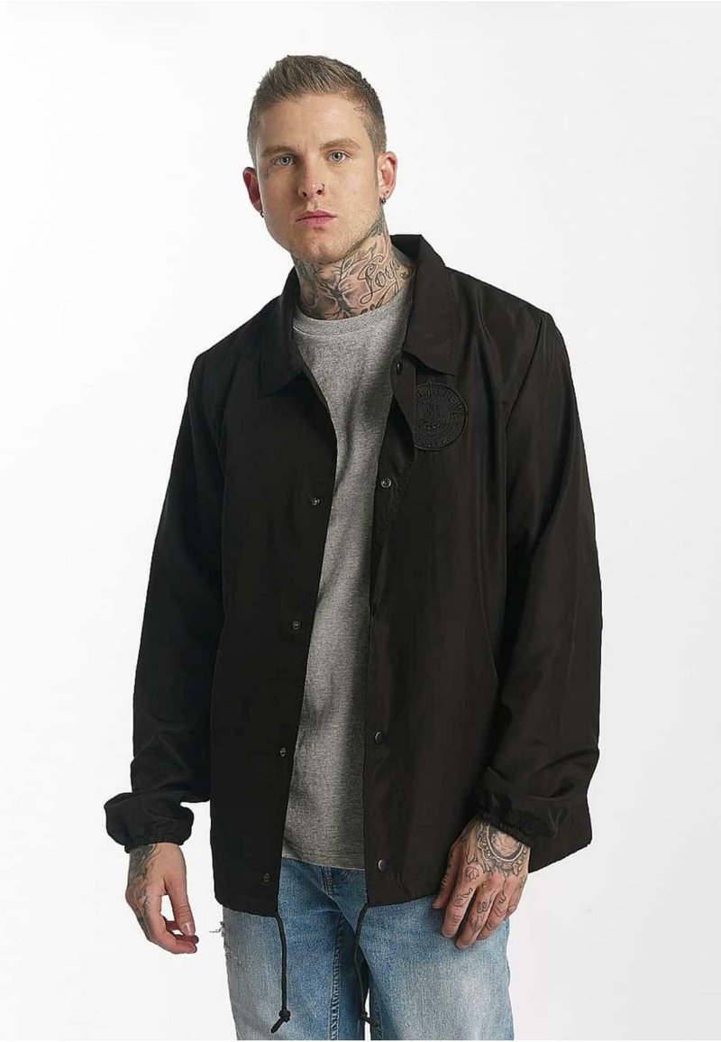 Czarna kurtka zapinana na guziki o fakturowej powierzchni, z okrągłą łatą na piersi, noszona na szarym T-shircie i niebieskich dżinsach.