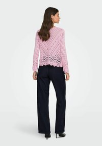 Femme debout de dos, portant un haut long à manches longues en tricot rose avec des détails de motif, un pantalon large de couleur foncée et des talons noirs.