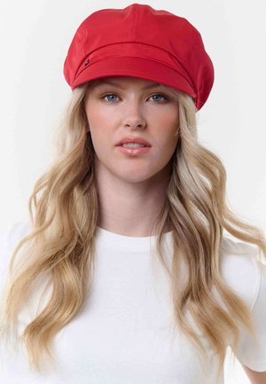 Blonde woman with loose waves wearing a bright red newsboy cap and white top, looking straight at the camera against a plain background.