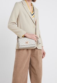 Crossbody bag in textured light gray leather with a decorative logo pattern, accented with gold hardware and a dual tassel zipper pull.