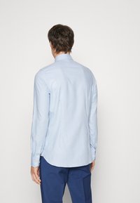 Light blue, long-sleeve shirt with a textured pattern, button cuffs, and a curved hem, displayed from the back. Tucked into navy pants.
