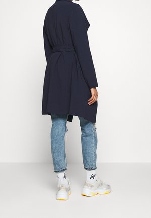 Persona que lleva un abrigo tipo trench azul marino con cinturón, vaqueros azul claro remangados, calcetines blancos con logo y zapatillas blancas con suelas amarillas, de pie sobre fondo blanco.