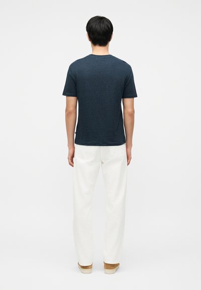 Άνδρας που στέκεται πλάτη, φορώντας σκούρο μπλε κοντομάνικο μπλουζάκι, λευκό παντελόνι και ανοιχτά καφέ παπούτσια μπροστά σε απλό λευκό φόντο.