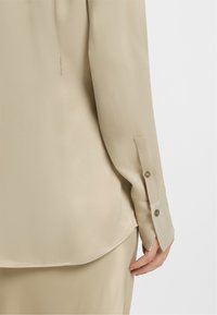 Beige satin long-sleeve shirt with buttoned cuffs worn over matching satin pants, showing part of a person’s hand and arm.