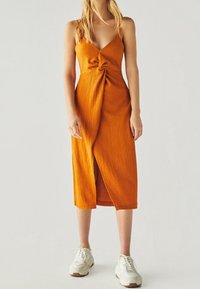 Knä-lång orange klänning med spaghettistroppar och en framknutning. Tillverkad av strukturerad tyg, med en sidoklyvning och bärs med vita sneakers.