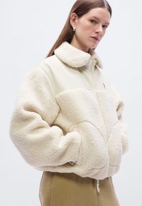 Μπεζ φλις μπουφάν με φερμουάρ μπροστά, ψηλό γιακά και μεγάλες τσέπες. Διαθέτει υφασμένο σχέδιο και μικρό λογότυπο στο στήθος.