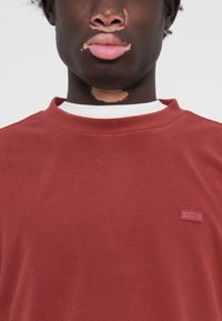 Sweatshirt en polaire bordeaux avec un col rond, tissu texturé, et une étiquette rectangulaire rouge cousue sur le côté gauche avec "RAINS" inscrit.