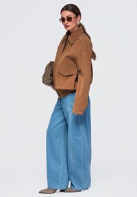 Femme portant une veste marron, un jean bleu large, des lunettes de soleil, tenant un sac à main marron, debout devant un fond clair uni.