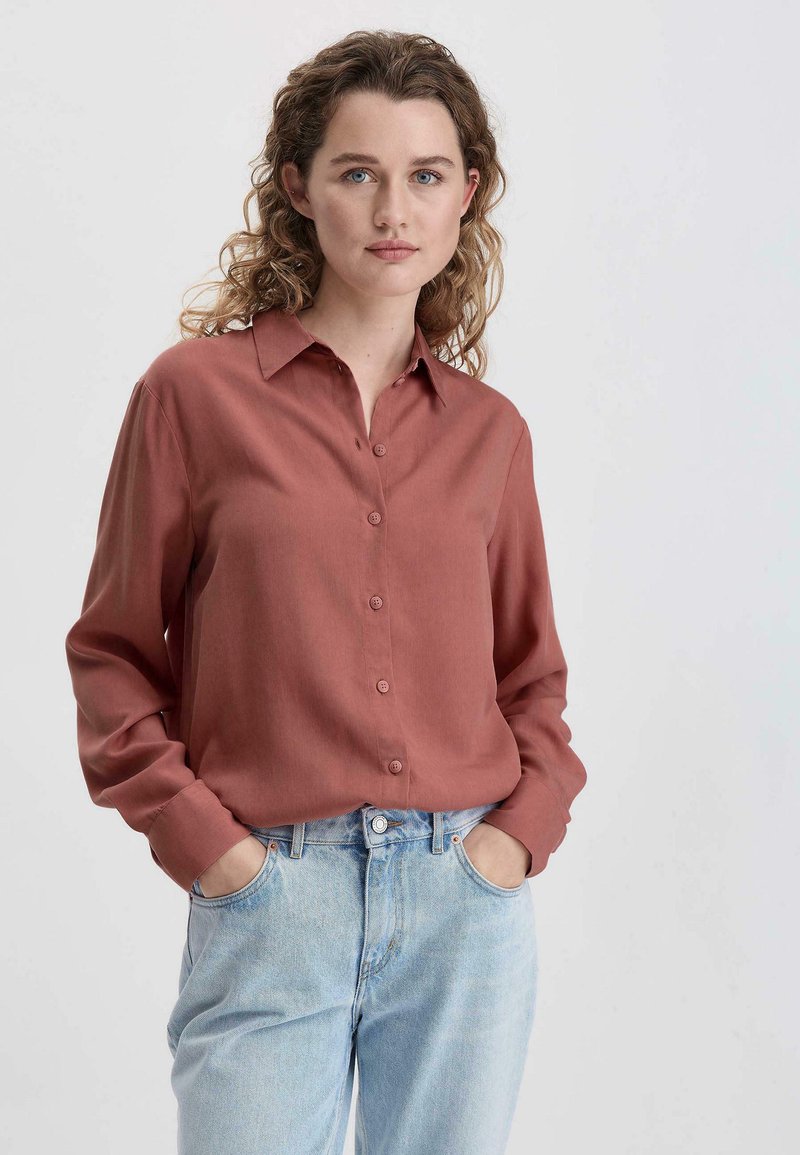 Rostfarbendes Hemd mit langen Ärmeln, glatter Textur und spitzen Kragen. Kombiniert mit hellblauen Jeans.