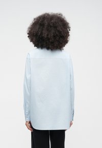 Person med mörkt lockigt hår bär en ljusblå långärmad tröja och svarta byxor, fotograferad bakifrån mot en enfärgad bakgrund.