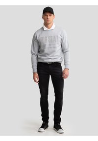 Mees, kes kannab halli dressipluusi tekstiga "BIG STAR", valget kraega särki, musti teksapükse, musta nokamütsi ja musti tossusid, seisab halli tausta ees.