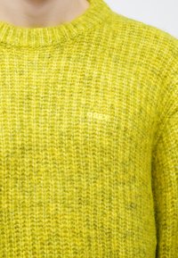 Hele kollane soonikkoeline kootud kampsun, millel on ümar kaelus ja väike samas toonis tikitud "OBEY" logo.