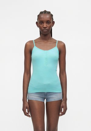 Donna con i capelli intrecciati che indossa una canottiera celeste con bottoni e pantaloncini di jeans, in piedi con le braccia rilassate lungo i fianchi, sfondo neutro.