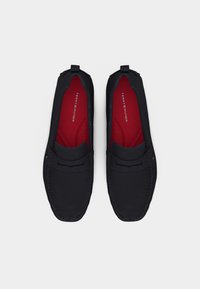 Ζευγάρι μαύρα μοκασίνια με κόκκινες εσωτερικές σόλες, με την επωνυμία "Tommy Hilfiger", εμφανίζεται από πάνω σε λευκό φόντο.