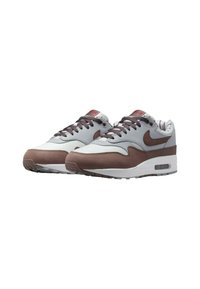 Sneakers Nike Air Max 1 charakteryzują się szaro-brązową skórą, wyściełanym kołnierzem, misternym przeszyciem oraz widoczną jednostką amortyzacji powietrznej w podeszwie.