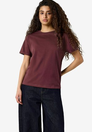 Ung kvinde med langt krøllet hår iført en ensfarvet vinrød t-shirt og mørkeblå vidde bukser, stående med den ene hånd på hoften.